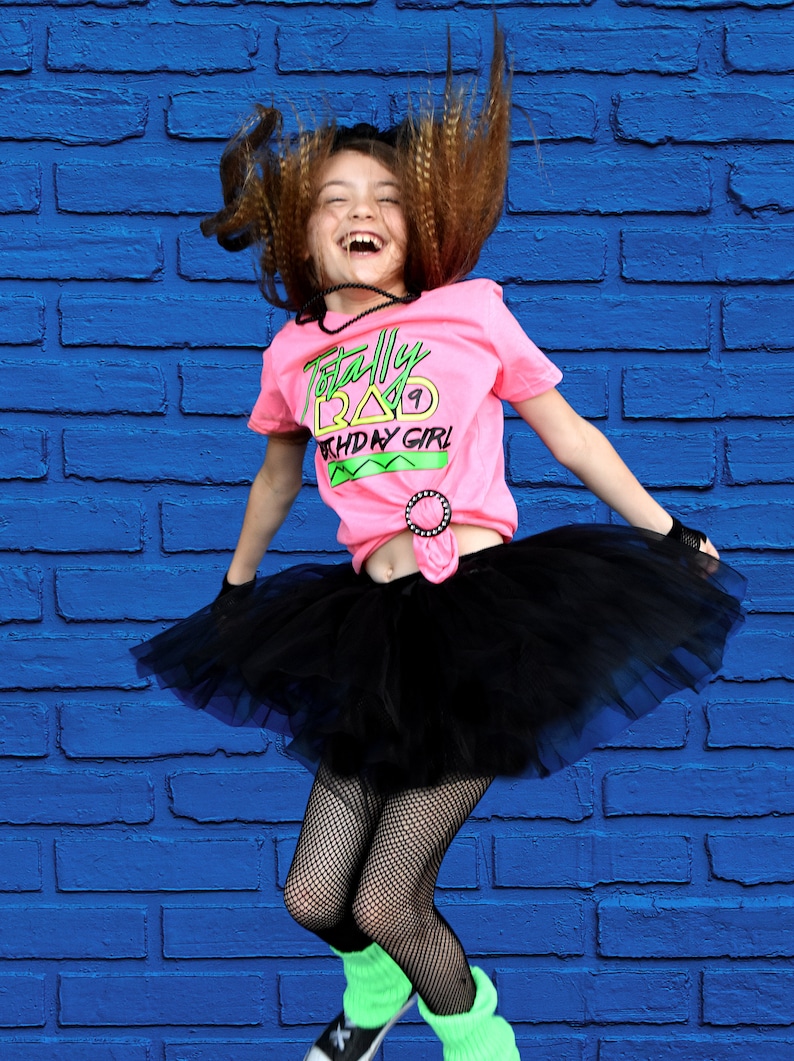 May include: A girl wearing a pink t-shirt with the text "Totally Rad Birthday Girl", a black tutu, fishnet stockings, and green leg warmers. Her hair is styled in a retro fashion, and she is jumping in front of a blue brick wall.