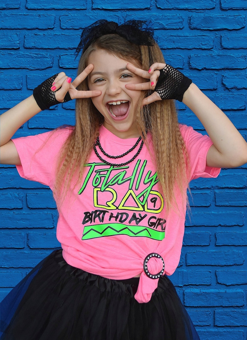 May include: A pink t-shirt with the text "Totally Rad Birthday Girl" in green and yellow, paired with a black tutu and fishnet gloves. The outfit is styled in an 80s theme, with a black bow and beaded necklace.