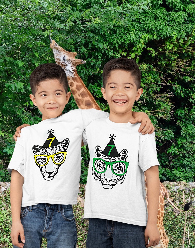 May include: Two boys wearing white t-shirts with animal face graphics and the number 7 on party hats. One shirt features a leopard with yellow glasses, the other a tiger with green glasses. Both are smiling in front of a green background.