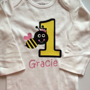 May include: White baby bodysuit with long sleeves, featuring a yellow number 1 and a cartoon bee. The bee is black and yellow with a pink heart-shaped wing and a small crown. The name "Gracie" is printed in pink below the number.