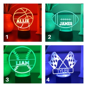 Puede incluir: Cuatro luces nocturnas iluminadas con tem&aacute;tica deportiva. Los dise&ntilde;os incluyen una pelota de baloncesto con "ALLIE", un bal&oacute;n de f&uacute;tbol americano con "JAMES", una pelota de b&eacute;isbol con "LIAM" y banderas a cuadros cruzadas con "TYLER". Cada luz se asienta sobre una base negra.
