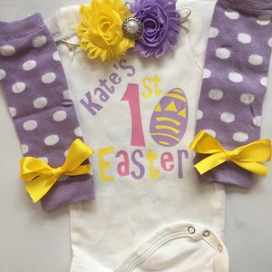 May include: A white baby onesie with the text "Kate's 1st Easter" and an Easter egg graphic. The outfit includes purple polka dot leg warmers with yellow bows and a floral headband with yellow and purple flowers.