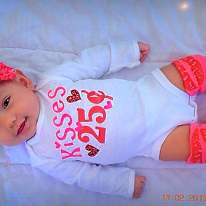 May include: A baby in a white onesie with pink and red text reading "Kisses 25p", a red floral headband, and pink leg warmers with red detailing and bows. The baby is lying on a white blanket.