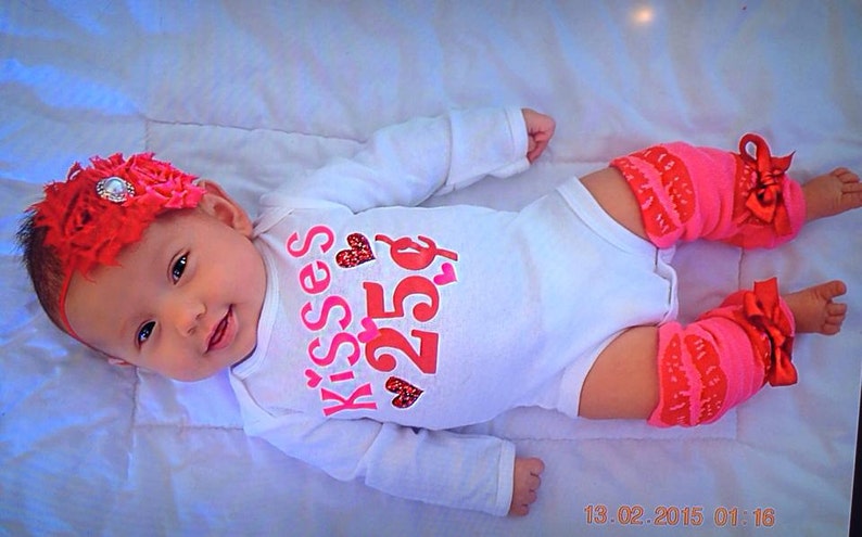 May include: A baby in a white onesie with pink and red text reading "Kisses 25p", a red floral headband, and pink leg warmers with red detailing and bows. The baby is lying on a white blanket.