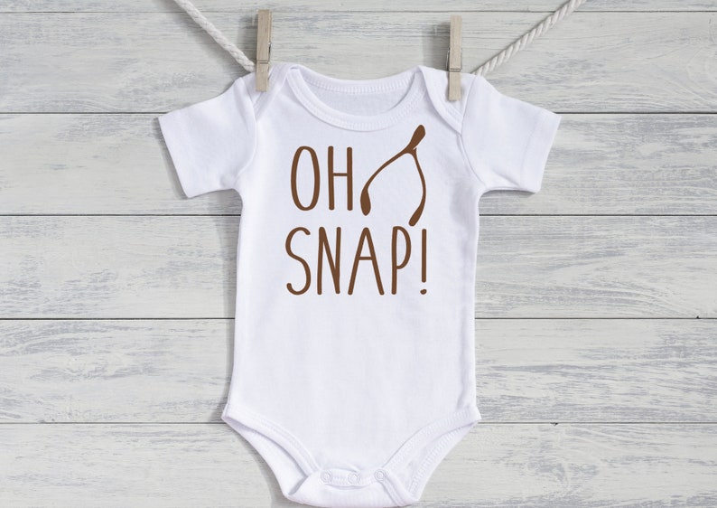 May include: White baby onesie with short sleeves, featuring the phrase "OH SNAP!" in brown lettering and a brown wishbone graphic. The onesie is hanging from a clothesline with a wooden clothespin.