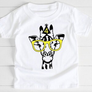 May include: White t-shirt featuring a black and yellow graphic of a giraffe wearing yellow glasses and a party hat with the number 4. The giraffe design is centered on the front of the shirt.
