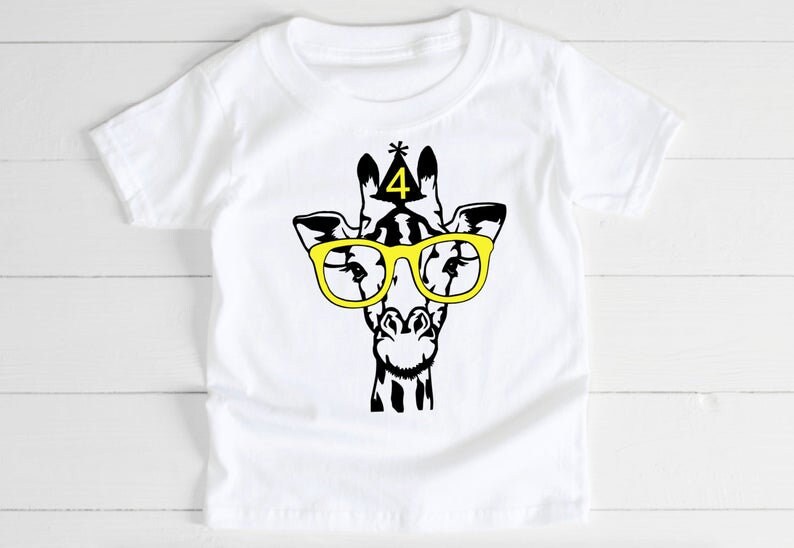 May include: White t-shirt featuring a black and yellow graphic of a giraffe wearing yellow glasses and a party hat with the number 4. The giraffe design is centered on the front of the shirt.