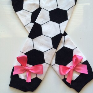 May include: A pair of soccer-themed leg warmers. The leg warmers are white with black soccer ball patterns and black cuffs. Each leg warmer has a bright pink bow attached near the ankle.