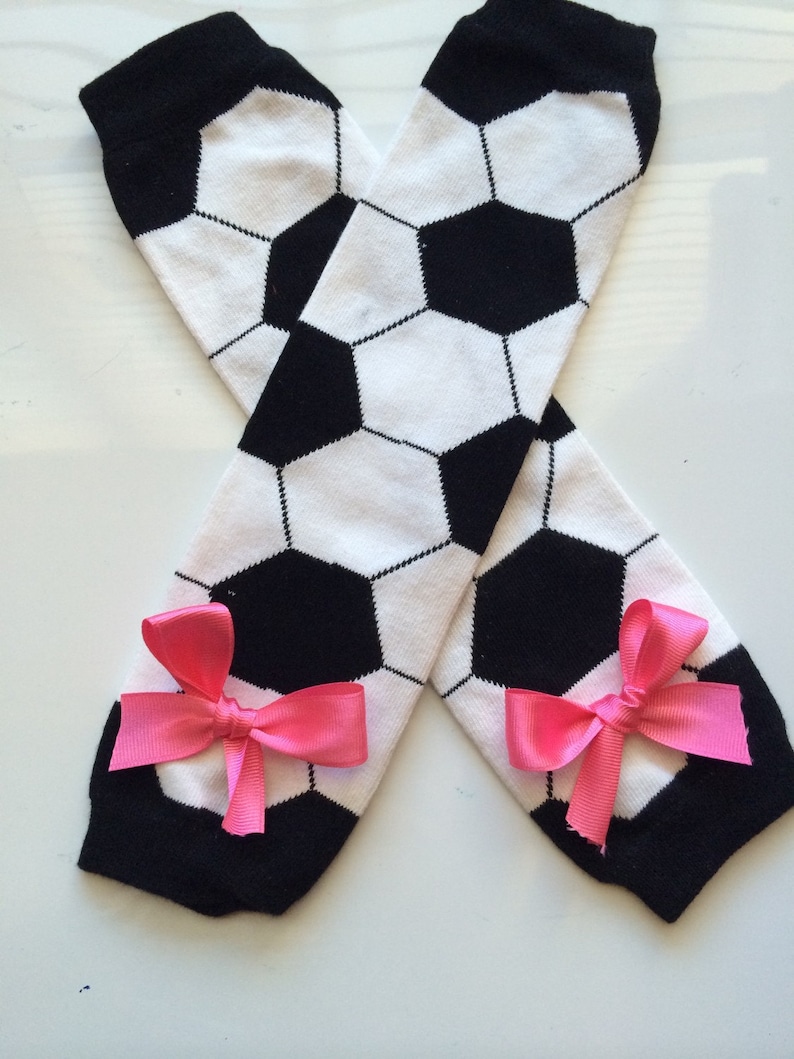 May include: A pair of soccer-themed leg warmers. The leg warmers are white with black soccer ball patterns and black cuffs. Each leg warmer has a bright pink bow attached near the ankle.