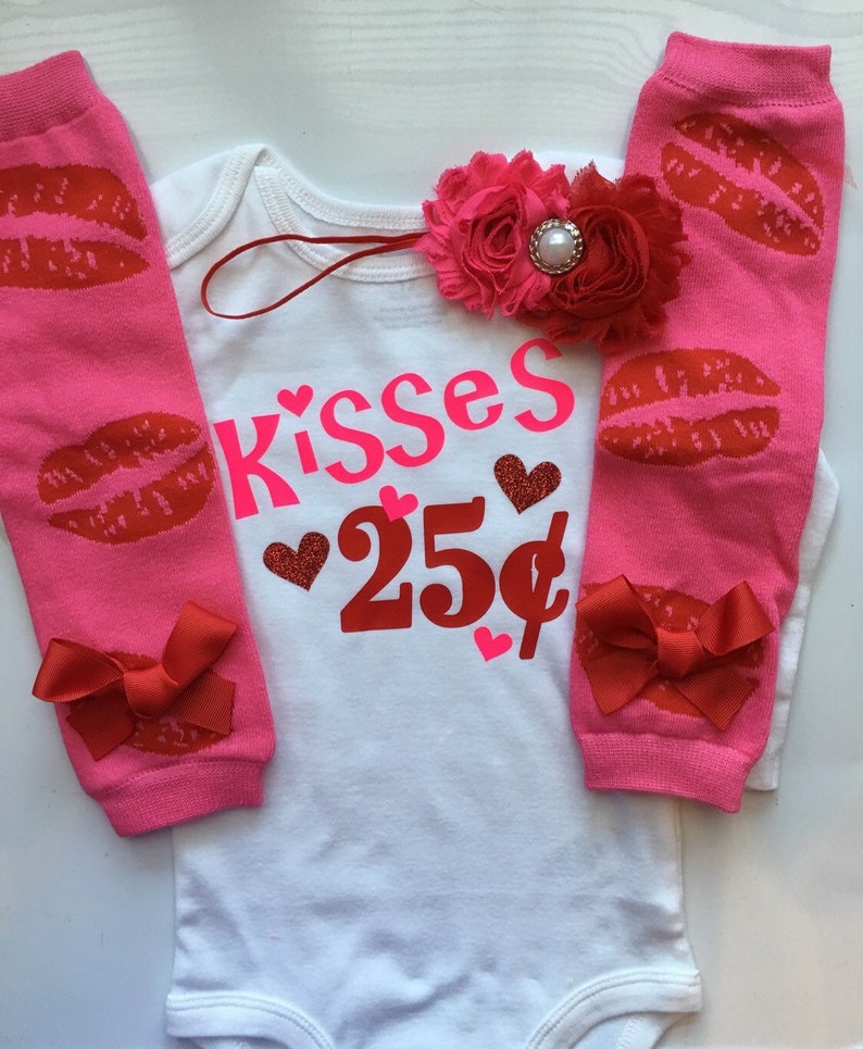 May include: A white baby bodysuit with the text "Kisses 25p" in pink and red, with red heart graphics. Pink leg warmers with red lip prints and red bows, plus a pink floral headband.