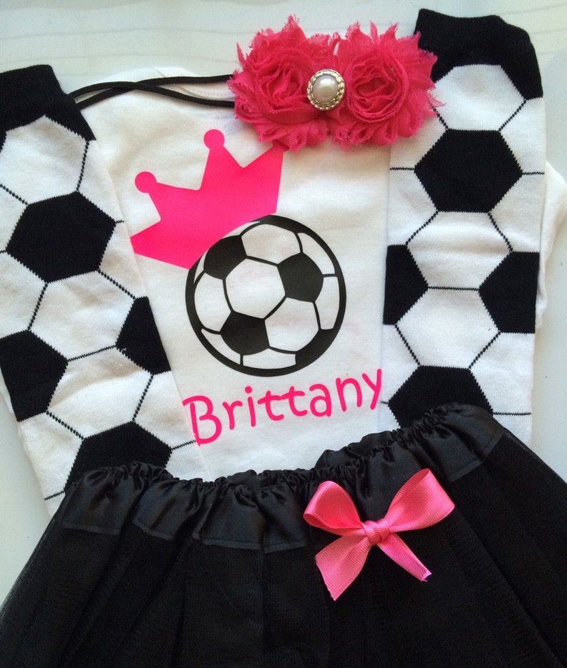 May include: A soccer-themed outfit featuring a white t-shirt with a soccer ball and crown graphic, the name "Brittany", and black and white soccer ball socks. A black tutu skirt with a pink bow and a pink floral headband complete the ensemble.