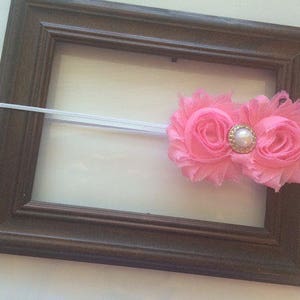 May include: A brown wooden picture frame with a white elastic band and a pink floral headband. The headband features two pink fabric flowers with a gold and pearl center. The frame is empty.