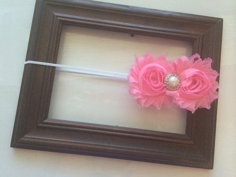 May include: A brown wooden picture frame with a white elastic band and a pink floral headband. The headband features two pink fabric flowers with a gold and pearl center. The frame is empty.