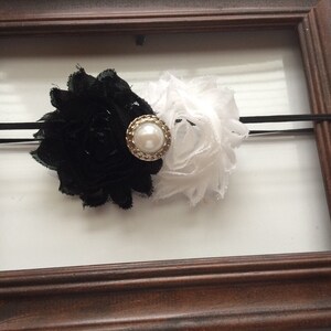 May include: A black and white floral headband with a pearl-centered embellishment. The headband features a black flower on one side and a white flower on the other, connected by a black elastic band.