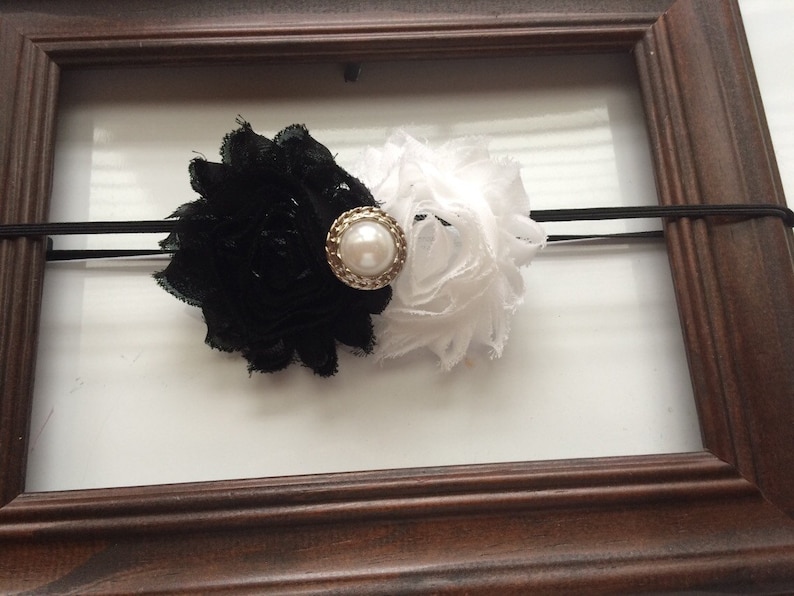 May include: A black and white floral headband with a pearl-centered embellishment. The headband features a black flower on one side and a white flower on the other, connected by a black elastic band.