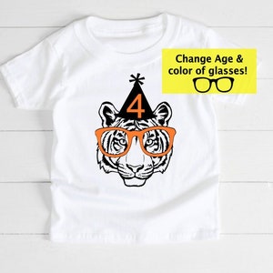 May include: White t-shirt featuring a tiger face wearing orange glasses and a black party hat with the number 4. The text "Change Age & color of glasses!" is in a yellow box. A fun birthday shirt.