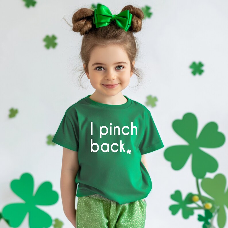 May include: A young girl wearing a green t-shirt with the text "I pinch back" in white. She has a green bow in her hair and is wearing green pants. The background features shamrock decorations.