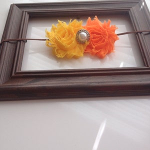 May include: A handmade headband featuring two fabric flowers in yellow and orange, connected by a thin brown cord. A pearl-centered embellishment sits between the flowers. The headband is displayed inside a brown wooden picture frame.