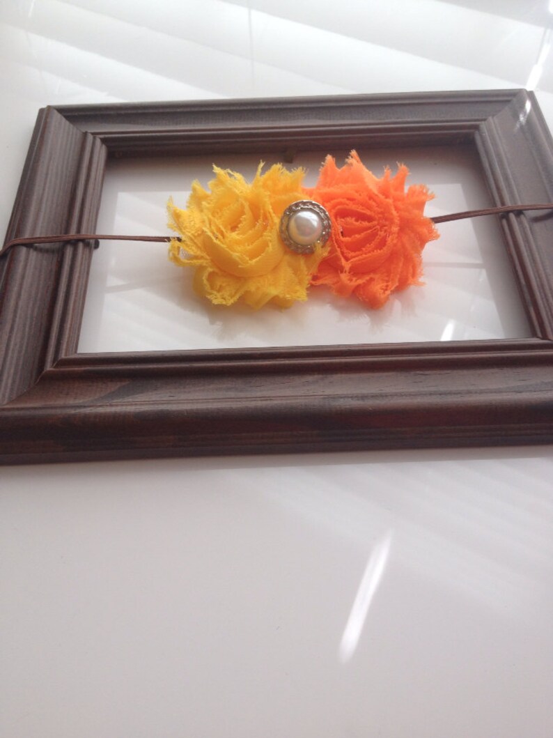 May include: A handmade headband featuring two fabric flowers in yellow and orange, connected by a thin brown cord. A pearl-centered embellishment sits between the flowers. The headband is displayed inside a brown wooden picture frame.