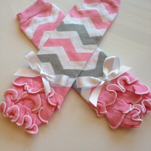 May include: Pink and white chevron patterned leg warmers with gray accents. The leg warmers have a pink ruffled trim and are tied with white satin bows. These baby leg warmers are a cute accessory.