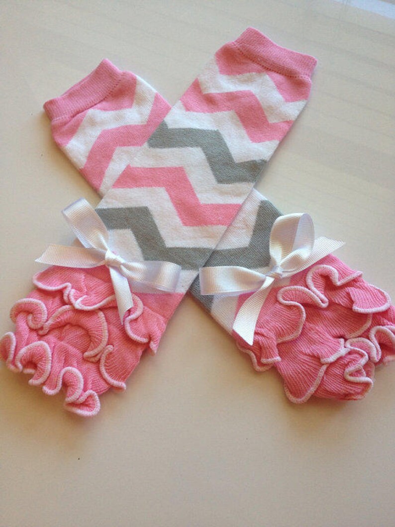 May include: Pink and white chevron patterned leg warmers with gray accents. The leg warmers have a pink ruffled trim and are tied with white satin bows. These baby leg warmers are a cute accessory.