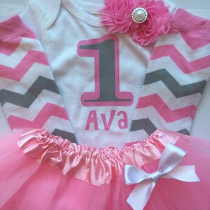May include: A white babygrow with pink and grey chevron sleeves, featuring a large grey number "1" outlined in pink, and the name "Ava" in pink. A pink tutu skirt with a white bow and a pink flower embellishment.