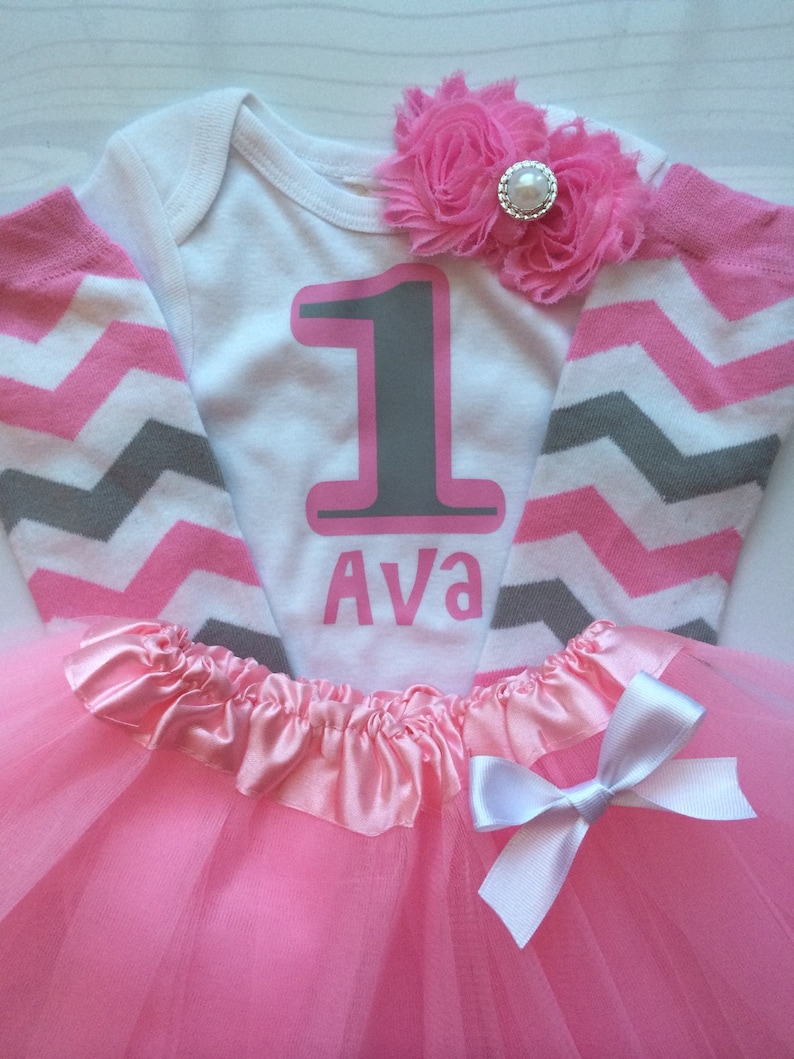 May include: A white babygrow with pink and grey chevron sleeves, featuring a large grey number "1" outlined in pink, and the name "Ava" in pink. A pink tutu skirt with a white bow and a pink flower embellishment.