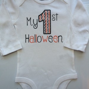 May include: White long-sleeved baby bodysuit with the text "My 1st Halloween" in black and orange. The number "1" is black with an orange and white zig-zag pattern. A festive outfit for a baby's first Halloween.