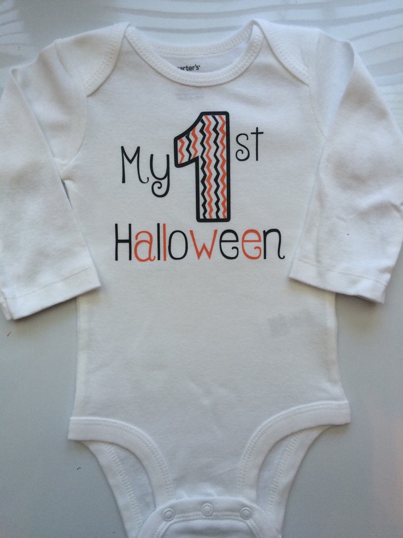 May include: White long-sleeved baby bodysuit with the text "My 1st Halloween" in black and orange. The number "1" is black with an orange and white zig-zag pattern. A festive outfit for a baby's first Halloween.