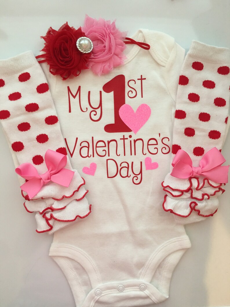K&ouml;nnte beinhalten: Ein wei&szlig;er Baby-Body mit dem Text "My 1st Valentine's Day" in Rot, einem rosa Herz und rosa Herzen. Das Outfit umfasst rot gepunktete Beinw&auml;rmer mit rosa Schleifen und ein rot-rosa Blumenstirnband.