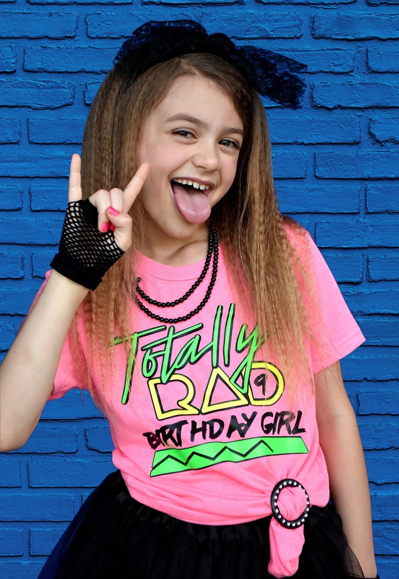 May include: A girl in a pink t-shirt with the words "Totally Rad Birthday Girl" in neon green and yellow. She wears a black net glove, a black beaded necklace, and a black bow. The background is a blue brick wall.