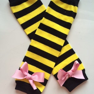 May include: Leg warmers in a yellow and black striped pattern. The leg warmers have black cuffs and are decorated with pink bows. These leg warmers are designed to be worn on the legs. The stripes are horizontal.