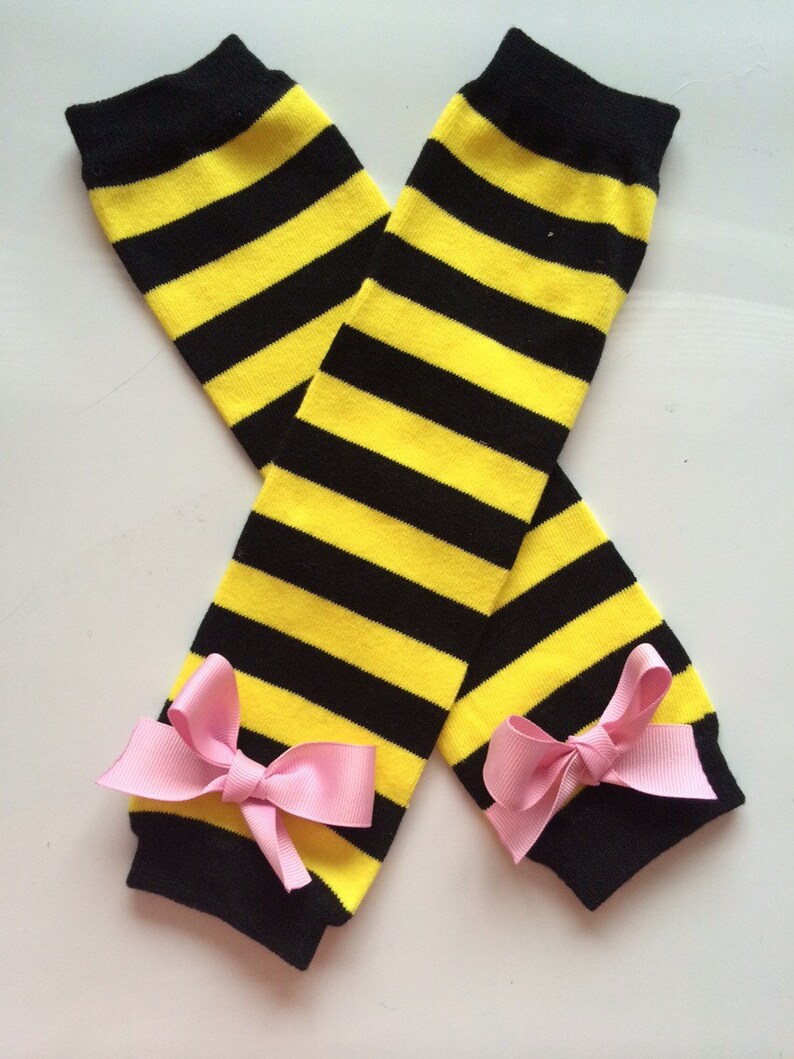 May include: Leg warmers in a yellow and black striped pattern. The leg warmers have black cuffs and are decorated with pink bows. These leg warmers are designed to be worn on the legs. The stripes are horizontal.