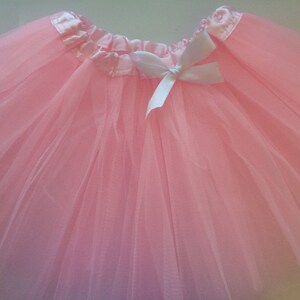 May include: A pale pink tutu skirt, constructed from multiple layers of tulle. The waistband is a satin ribbon, adorned with a white bow. Ideal for dance, fancy dress, or special events.