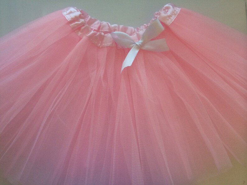 May include: A pale pink tutu skirt, constructed from multiple layers of tulle. The waistband is a satin ribbon, adorned with a white bow. Ideal for dance, fancy dress, or special events.