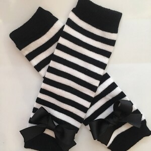 May include: A pair of black and white striped leg warmers with black bows at the ankles. The leg warmers have a thick black band at the top and are made of a soft, knitted material. The bows add a decorative touch.
