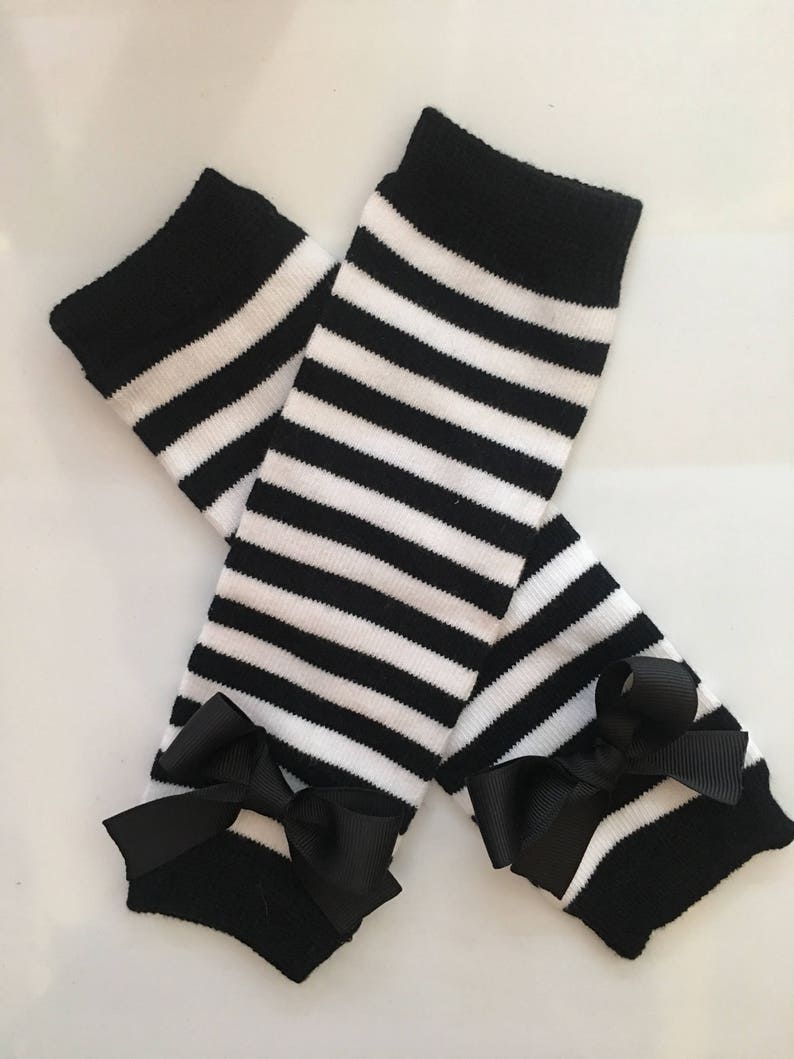 May include: A pair of black and white striped leg warmers with black bows at the ankles. The leg warmers have a thick black band at the top and are made of a soft, knitted material. The bows add a decorative touch.