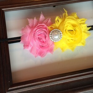 May include: A pink and yellow floral headband with a pearl and rhinestone centre. The headband is made of a black elastic band and is set against a white background within a brown wooden frame.
