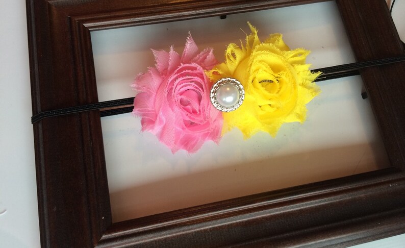 May include: A pink and yellow floral headband with a pearl and rhinestone centre. The headband is made of a black elastic band and is set against a white background within a brown wooden frame.