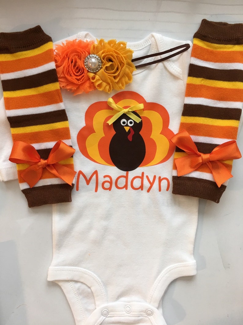 May include: A white baby bodysuit with a turkey graphic and the name "Maddyn" in orange. The outfit includes striped leg warmers with orange bows and a floral headband with a pearl accent. The colors are brown, orange, yellow, and white.