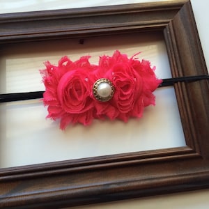 May include: A bright pink fabric flower headband with a pearl and gold-tone center. The headband is set against a brown wooden picture frame with a black elastic band.