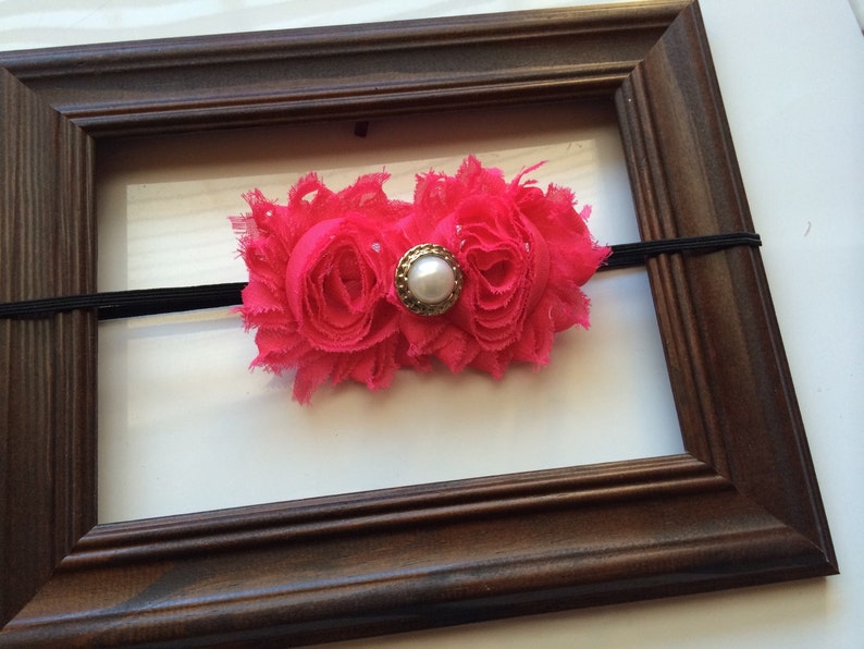 May include: A bright pink fabric flower headband with a pearl and gold-tone center. The headband is set against a brown wooden picture frame with a black elastic band.