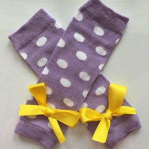 May include: A pair of lavender leg warmers with white polka dots and yellow bows. The leg warmers are made of a soft knit fabric and are designed to be worn on the legs. The bows are made of a satin ribbon.
