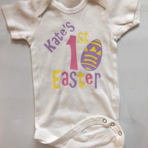 May include: White baby onesie with the text "Kate's 1st Easter". The text is in purple, pink, and yellow. An Easter egg graphic is also on the onesie. The onesie has short sleeves and snaps at the bottom.