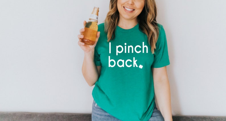 May include: A green t-shirt with the text "I pinch back" in white, along with a small shamrock. The person is holding a beer bottle. The shirt is a solid color and has a round neckline.