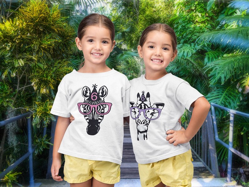 May include: Two young girls wearing white t-shirts with animal graphics. One shirt features a zebra wearing pink glasses and a party hat with the number 6. The other shirt has a giraffe with purple glasses and a party hat with the number 6.