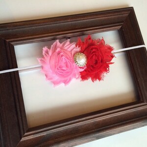 K&ouml;nnte beinhalten: Ein Baby-Stirnband mit zwei Stoffblumen, eine rosa und eine rote, mit einem Perlen- und Strass-Mittelst&uuml;ck. Die Blumen sind an einem wei&szlig;en Gummiband befestigt. Das Stirnband wird in einem dunkelbraunen Holzrahmen pr&auml;sentiert.