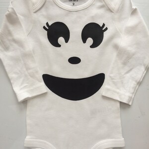 May include: White baby onesie with a black ghost face design. The design includes large eyes with eyelashes, a small nose, and a wide smile. The onesie has long sleeves and the text "carter's 9" is printed on the neckline.