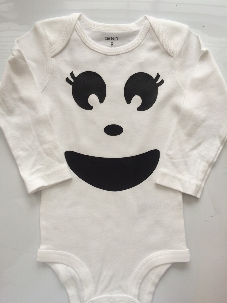 May include: White baby onesie with a black ghost face design. The design includes large eyes with eyelashes, a small nose, and a wide smile. The onesie has long sleeves and the text "carter's 9" is printed on the neckline.