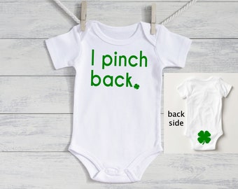 May include: White baby bodysuit with green text that reads "I pinch back." A small green shamrock is next to the word "back." The back of the bodysuit has a green shamrock. The bodysuit is hanging on a clothesline.